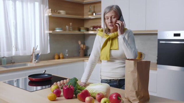A woman and an elderly woman converse on cell phones in kitchens