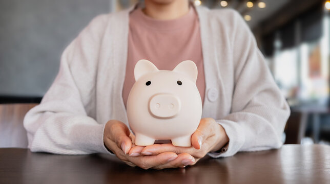 Woman hands holding cute white piggy bank on wooden table. Symbolizing saving money, financial planning, investment and budget.