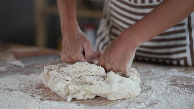 Hands of professional baker in maroon apron(white uniform) kneading dough on floury wooden table, filled with baking tools and ingredients, in artisan bakery shop 