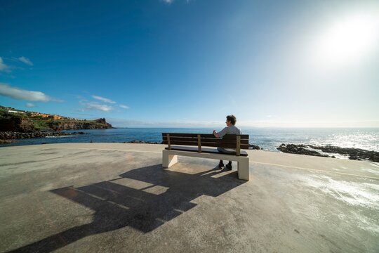 A 60-70 year old woman sits on a bench, waterfront on the south coast with the Atlantic Ocean behind it, Madeira, Portugal