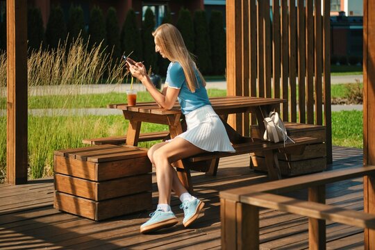 Active woman sits in wooden gazebo in an outdoor area and checks online schedule of classes at sports club using mobile internet of her smartphone