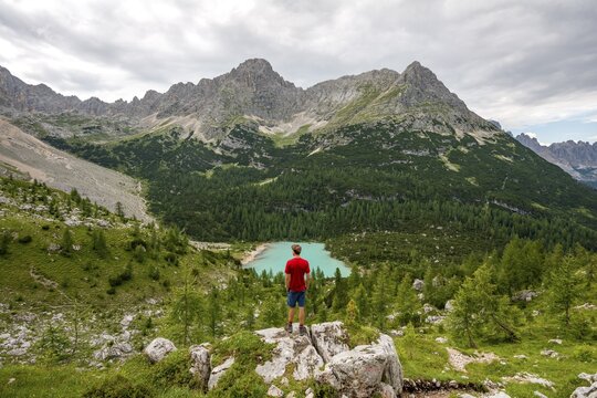 Young man, hiker stands on rocks and looks at turquoise green Sorapiss lake and mountain landscape, Dolomites, Belluno, Italy
