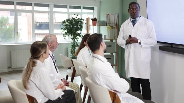 During lesson, African male doctor with remote controller tell material, speaks to group of students. Lecturer leads class in audience, explains material to audience, provide practical cases 