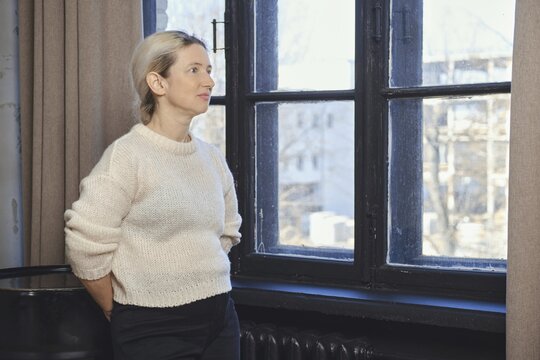 A woman stands next to a large window, lost in thought as she gazes outside. The sunlight filters into the room, highlighting her soft sweater and the serene atmosphere