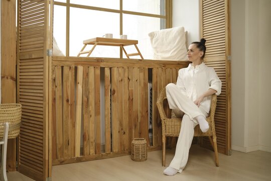 A woman in white attire sits gracefully on a minimalist wicker chair, bathed in soft natural light. The contemporary interior exudes calmness and simplicity, creating a serene atmosphere