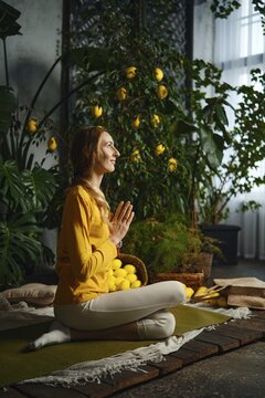 A woman sits on a mat with her legs crossed holding hands in a prayer position. She enjoys yoga in a bright room with many lemon plants around her. The scene is full of plants and natural light