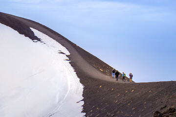 Group of hikers with trekking poles walk along slope of Mount Etna beneath clear blue sky. Concept of collective determination and shared challenge of climbing dramatic volcanic mountain in Sicily. © Ina