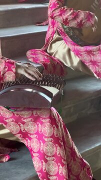 A woman in a pink traditional dress plays a musical instrument on stairs. The setting is serene and cultural, highlighting the beauty of music and tradition.