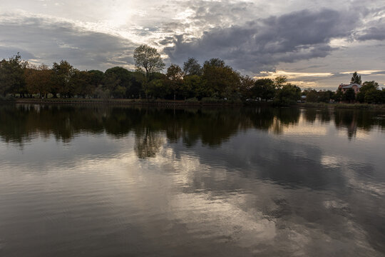 Suggestivo paesaggio lacustre al tramonto con nuvole drammatiche che si riflettono perfettamente sullo specchio d'acqua calmo, circondato da alberi e vegetazione rigogliosa in un'atmosfera serena.