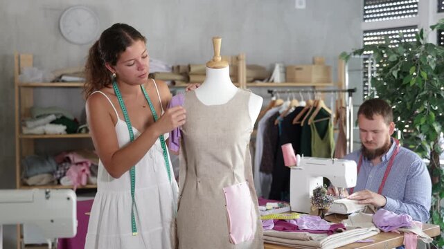 Enthusiastic young tailor stitching custom garment from lilac cotton jersey fabric on sewing machine in workshop, while female colleague adjusting sample dress on mannequin 