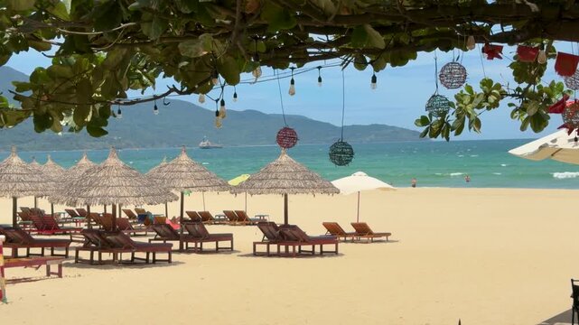 Da Nang beach cafe view framed by hanging lanterns and leaves, with thatched umbrellas, loungers, and mountains beyond the sea
