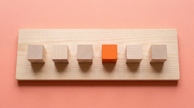 One orange wooden cube standing out among identical beige cubes on light wood board symbolizing uniqueness or individuality concept