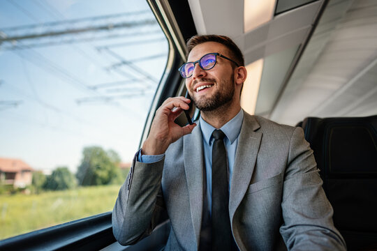 Smiling professional in suit looking out the window while holding a smartphone conversation on a modern passenger railway journey.