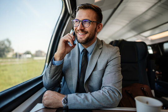 Smiling professional in suit looking out the window while holding a smartphone conversation on a modern passenger railway journey.