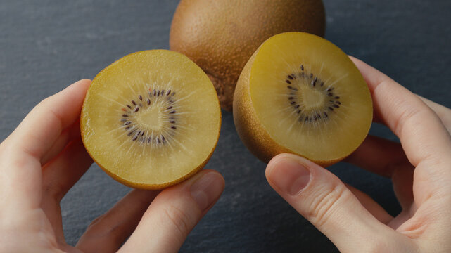 Close-up of female hands holding two halves of a ripe golden kiwi, both showing a heart-shaped core on dark gray background with blurred whole kiwi for healthy eating, gourmet food, culinary content.