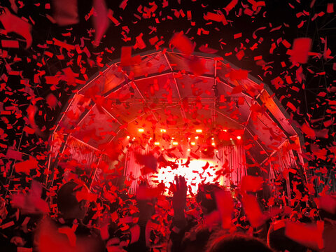 Multid&atilde;o euf&oacute;rica em festival noturno a celebrar sob ilumina&ccedil;&atilde;o vermelha vibrante e chuva de confetis em frente ao palco principal