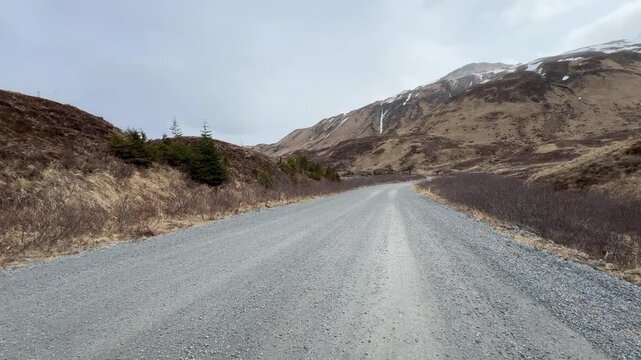 Tracking center-lane shot of a wide dirt pass road curving through a valley toward snowy peaks in Unalaska Island.
