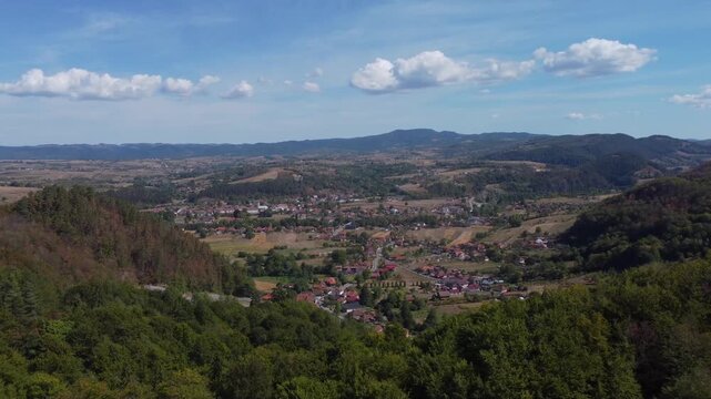 High aerial panorama of Bratca village in Romania, nestled among lush forested hills and rolling green countryside under a bright blue sky filled with fluffy white clouds.