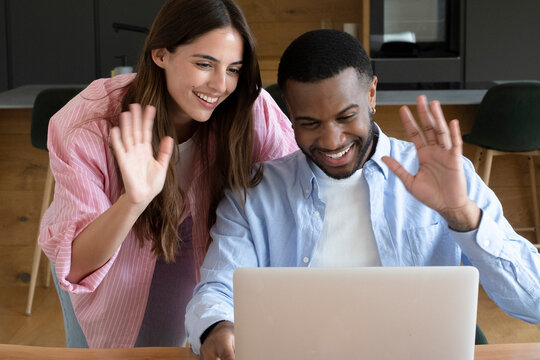 Young couple smiling and waving at a laptop during a joyful video call at home, sharing a virtual conversation, warmth and connection while catching up with friends and family.