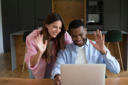 Young couple smiling and waving during a joyful video call on their laptop at home, connecting with family or friends in a warm conversation, enjoying a remote connection together.