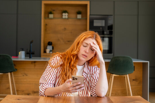 Redheaded woman at home kitchen table looking stressed and frustrated while reading her smartphone, holding her forehead in a kitchen, showing worry, fatigue and distraction