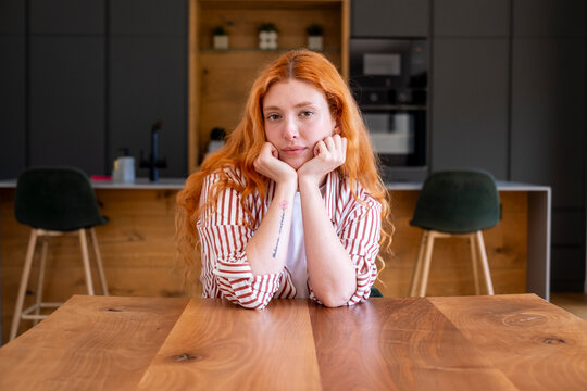 Redheaded woman leans on hands at wooden table in modern kitchen, wearing striped shirt and a thoughtful expression, portrait conveying calm, introspection and relaxed mood