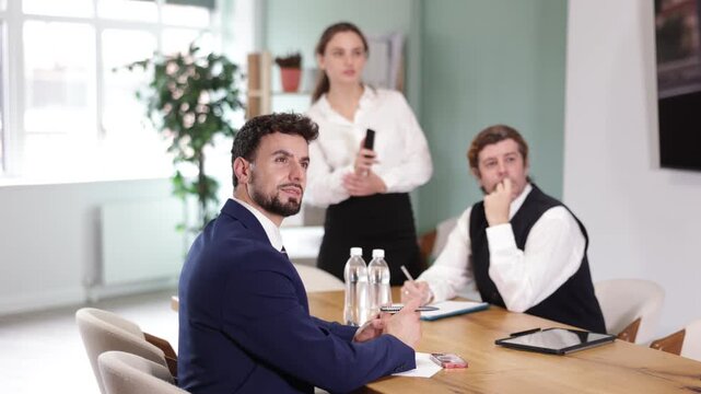Representatives of business professional community sitting at table and chatting, talking. Colleague with remote control telling at background, assist and helps manage presentation 