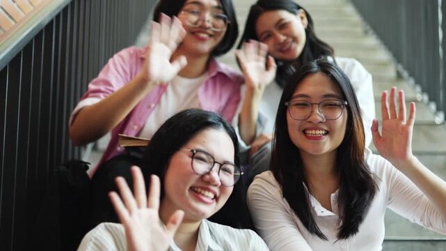 Group cheerful asian male and female students carrying backpacks and textbooks sit on the steps academic building smile and wave camera university building ready university studies.