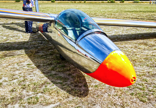 Chrome glider fuselage with orange nose cone on a grassy airfield, reflecting the sky and a person standing nearby