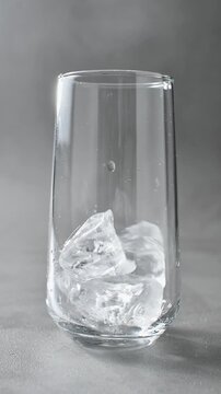 Ice cubes fall one by one into clear drinking glass on gray background in vertical studio shot.