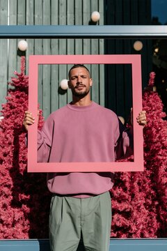 Man holding a square pink frame in front of pink trees and window for poster frame mockup and social media banner background
