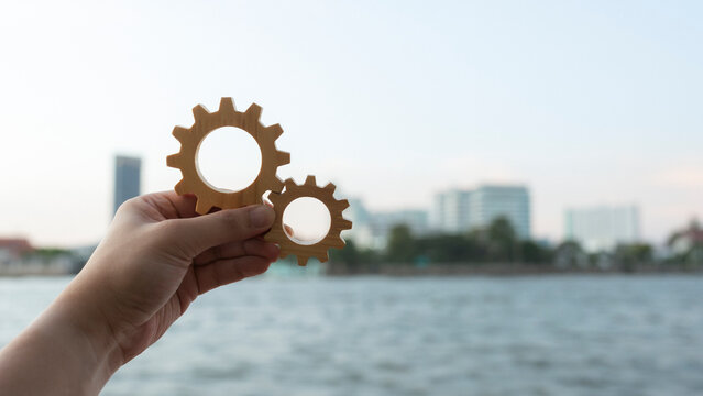 Business mechanism and engineering synergy concept with hand holding interlocking wooden gears against river and city skyline background for corporate teamwork, operations and urban development.