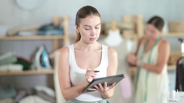 Careful young tailoress drawing dress model using tablet, other works with dummy in sewing studio 