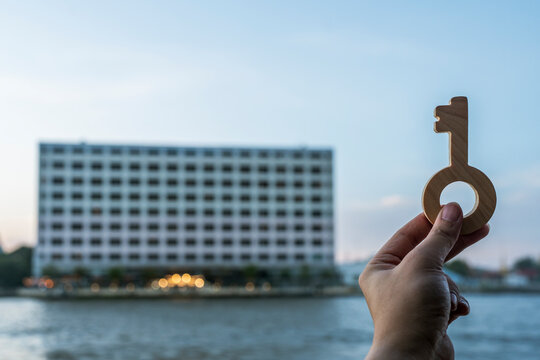 Real estate and apartment handover concept with hand holding wooden key against modern riverfront residential building for hotel room access, property investment and urban condo living.