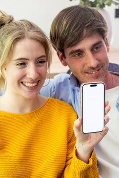 Young couple smiling and sharing a smartphone with a blank screen while sitting at home, showcasing technology, connection and happiness as they look at content on the phone.