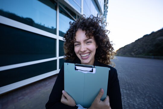Businesswoman smiling and holding folder outdoors near modern building