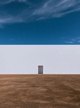 Empty field with a white wall in the background. the sky is a deep blue with a few wispy clouds scattered across it. the ground is a light brown color and appears to be dry and barren.