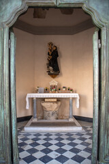 Antica cappella devozionale cattolica con altare in marmo e statua lignea sacra, incorniciata da un portale in legno rustico consumato dal tempo e pavimento a scacchi bianchi e neri. © Ilarialapreziosa
