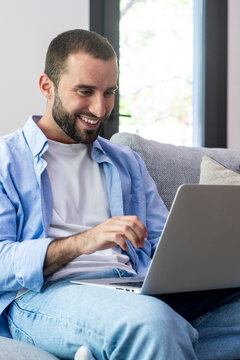 Young man working remotely on a laptop from his comfortable living room couch, smiling while browsing and multitasking, relaxed outfit conveying productivity, home everyday life.