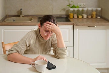 Worried woman in glasses sitting at kitchen table with cup and smartphone. Woman touching forehead...