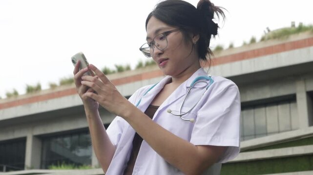Cheerful Asian female medical student walks in front of the academic building chatting happily on her smartphone classmate who is waiting inside the modern facility.