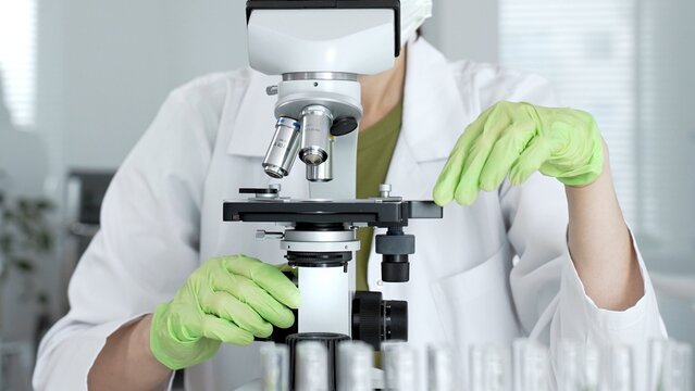 Female scientist wearing a lab coat and safety glasses working with a microscope, performing research and careful analysis in a modern laboratory for medical and scientific discovery