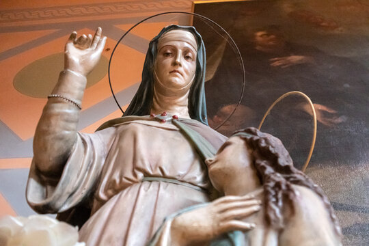Statua religiosa in gesso dipinto raffigurante una santa cattolica con velo e aureola sottile che compie un gesto di benedizione, situata all'interno di una chiesa storica decorata con affreschi.