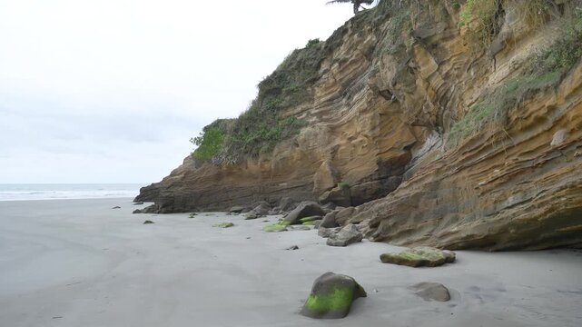 Striated sandstone cliff with moss covered rocks scattered on the wet sand at Mompiche beach, Esmeraldas Province, Ecuador, slow pan left handheld shot