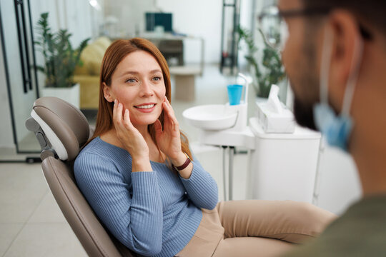 Woman smiling and touching her cheeks in a dental chair, discussing a treatment plan with the masked dentist, focusing on oral care and cosmetic dentistry consultation
