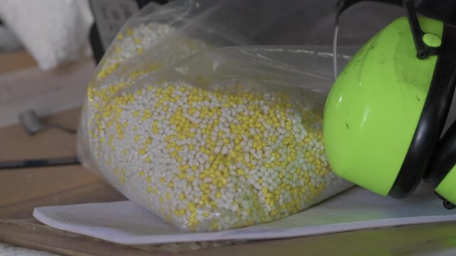 Hearing protection earmuffs left on a shelf next to a clear sample bag of white and yellow resin granules at a plastics processing plant