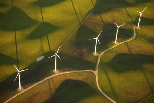 Aerial view of wind turbines standing in the rolling green hills of a wind farm during golden hour with long shadows stretching across the landscape in Rio Vista, California, United States.