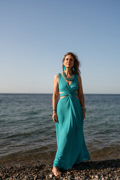 Woman beach turquoise. Elegant woman in flowing turquoise dress stands on a serene pebble beach with ocean waves.
