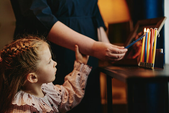 Caucasian child watching mother lighting Hanukkah menorah candles during Jewish holiday celebration at home. Image suitable for religious education campaigns