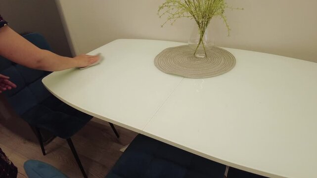 A person is wiping a white table in a dining area. The table is round with a woven mat placed on it. There are blue chairs around the table. The setting looks clean and organized. The activity shows 